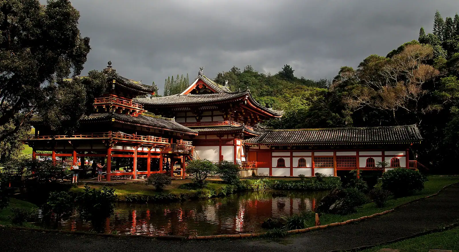 A red Japanese temple hall with wing corridors reflected in a still pond beneath green cliffs